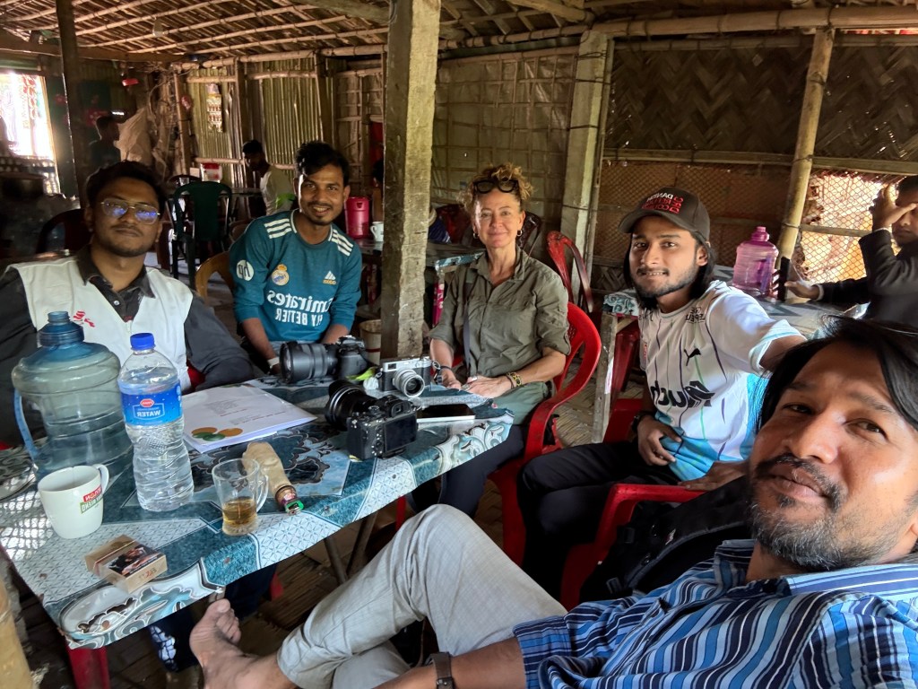 A group of five people gathered around a table in a rustic setting, with cameras, water bottles, and documents on the table. They appear to be engaged in a discussion.