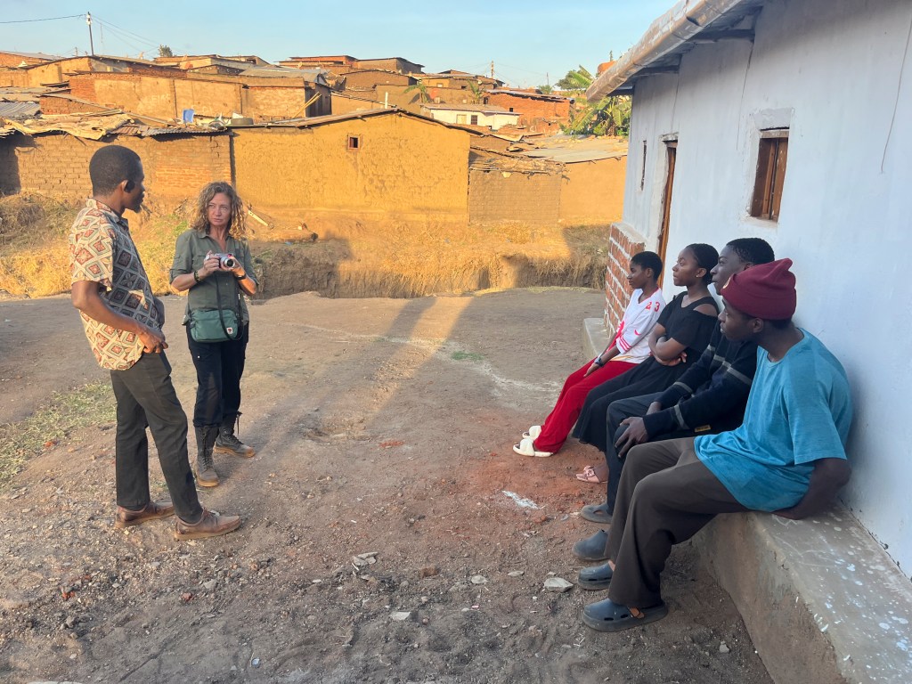 A woman holding a camera stands conversing with a man, while four young people sit on a wall in a rural area with mud houses in the background under a clear sky.