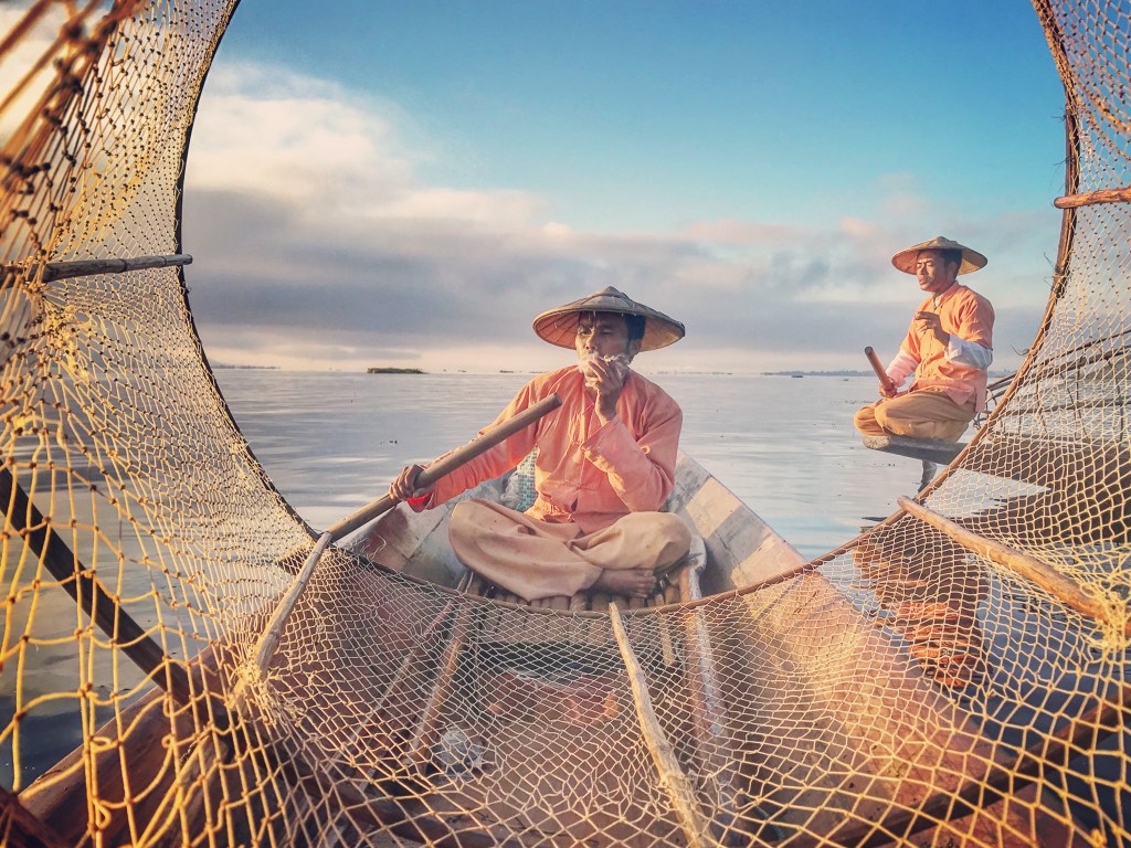 Two fishermen in traditional attire, one casting a net and the other sitting on a wooden boat, surrounded by calm water under a clear sky.