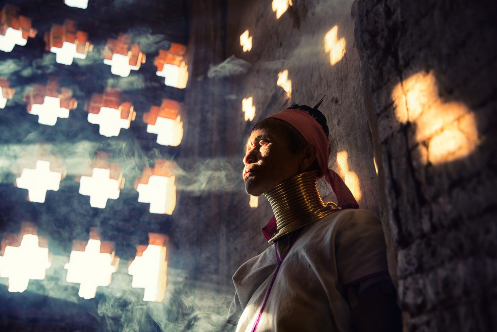 A woman with traditional brass neck rings stands in a dimly lit room, framed by patterned light coming through a window, with smoke creating a mystical atmosphere.