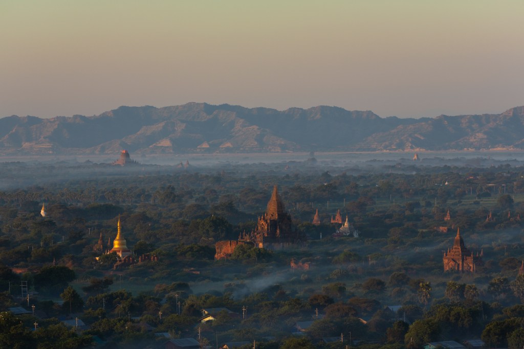 A panoramic view of ancient temples and pagodas in Bagan, Myanmar, surrounded by lush greenery and distant mountains during sunrise with mist.