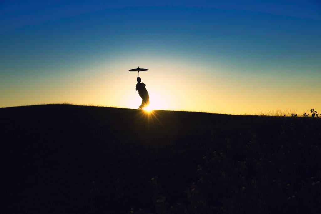Silhouette of a person holding an umbrella on a hilltop at sunset, with a vibrant gradient sky in the background.