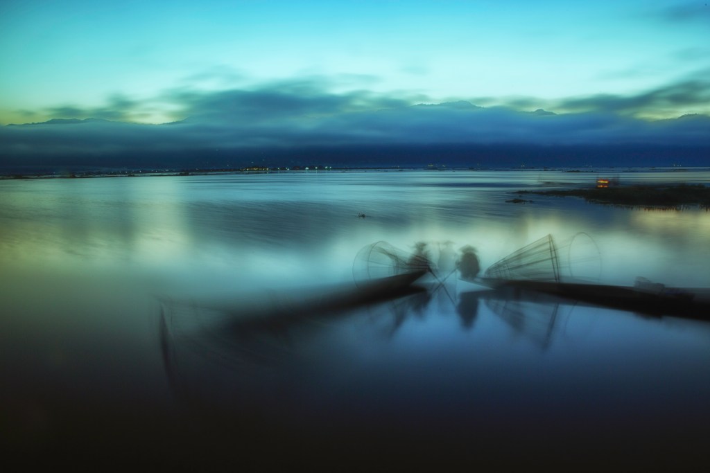A tranquil lake scene at dusk with soft blue tones, featuring fishermen in canoes silhouetted against a misty background.