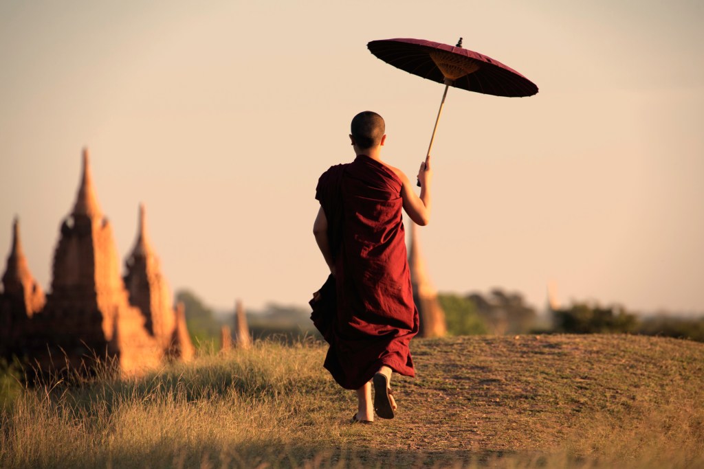 A monk in traditional robes walking on a grassy path, holding a large umbrella, with ancient temple structures in the background during sunset.