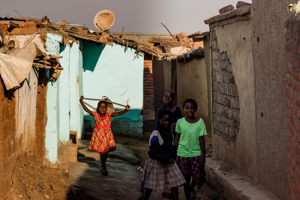 A group of children walking through a narrow alley in a residential area, with one girl in a red dress carrying a stick, while others are casually dressed in various colored shirts and skirts.