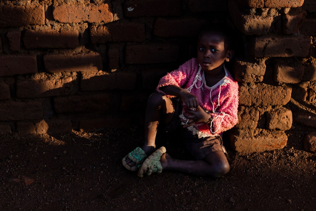 A child sitting against a brick wall, wearing a pink sweater and brown shorts, with a serious expression in a dimly lit environment.