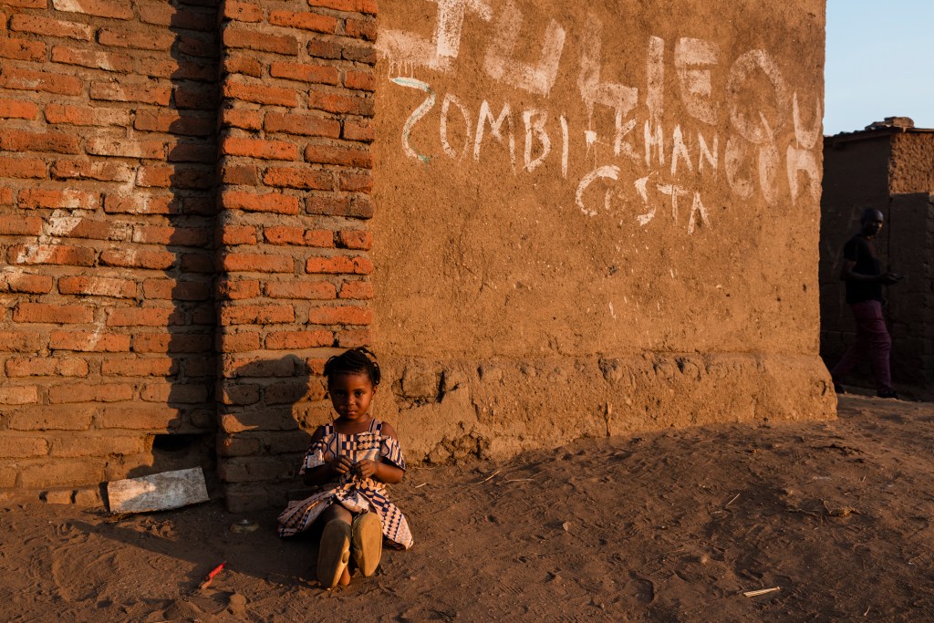 A young girl sitting on the ground beside a brick wall, wearing a patterned dress, with a sandy surface around her.