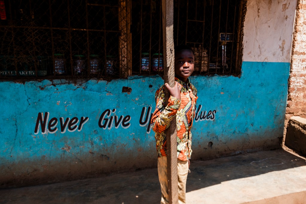 A young boy in a colorful patterned shirt stands beside a wooden post in front of a blue wall with the phrase 'Never Give Up' painted on it.