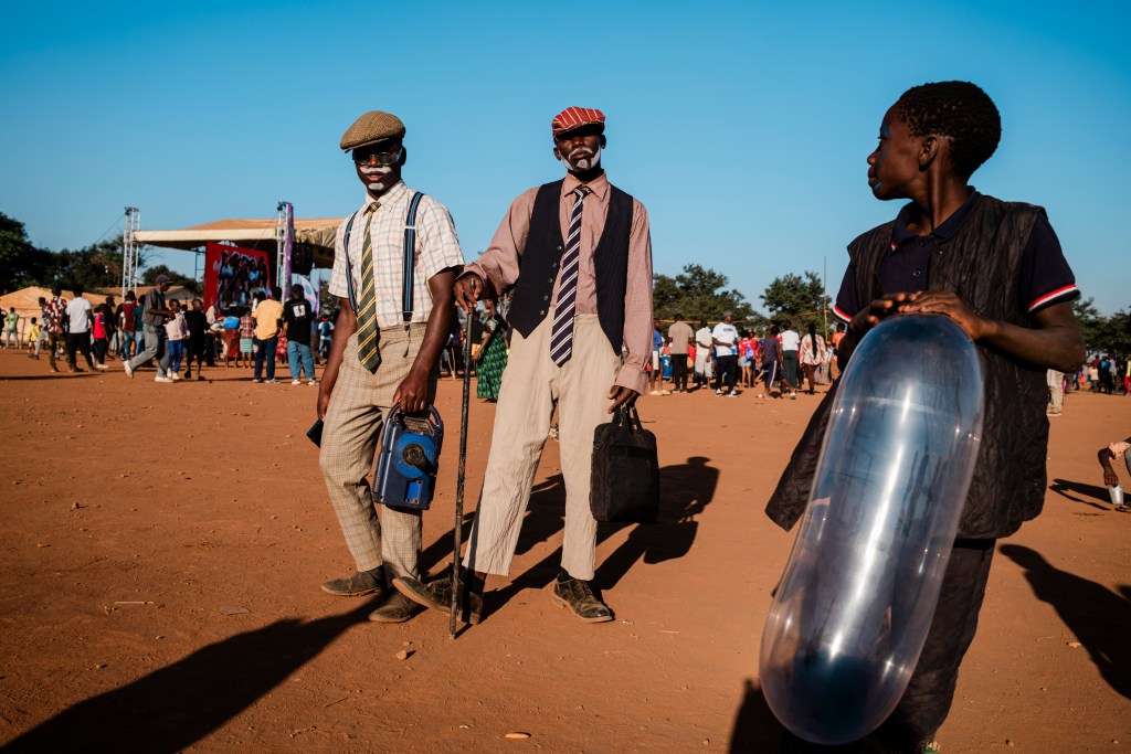 Two men dressed in vintage-style clothing stand in a crowd at an outdoor event, with a stage in the background. One man carries a briefcase and a cane, while the other holds a blue backpack. A young boy nearby is holding a large, transparent balloon.