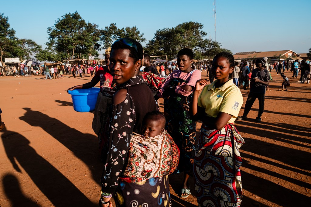 A group of women in traditional attire, with one woman carrying a baby on her back, standing in a bustling outdoor market setting. The background features a crowd and market stalls under a clear blue sky.