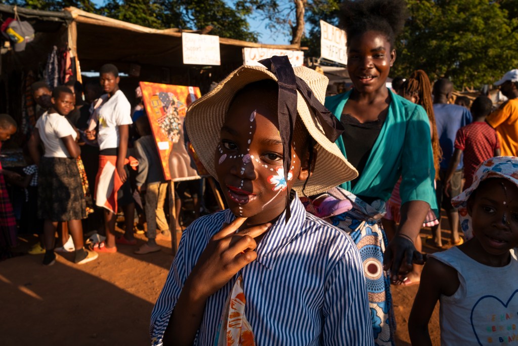 A young girl with face paint and a wide-brimmed hat poses for the camera in a busy outdoor market. People are gathered around various stalls in the background, with vibrant colors and artwork visible.