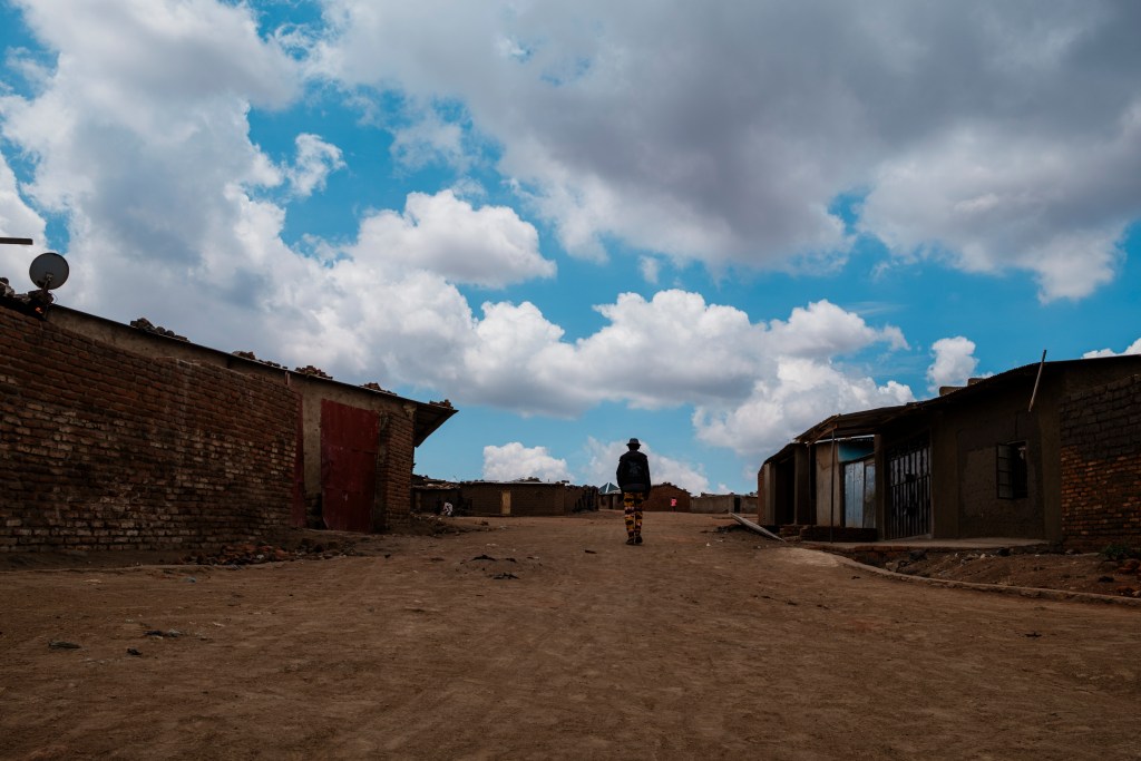 A person walking down a dusty road between brick and concrete buildings under a cloudy blue sky.